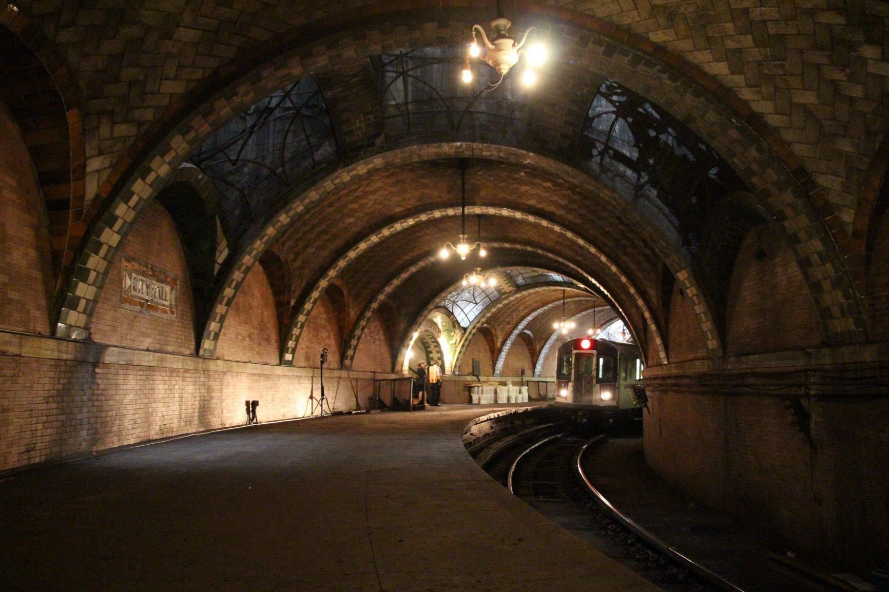 Inside what looks as an old subway stop with large archways and classic subway tile. A subway train car can be seen in the distance. 