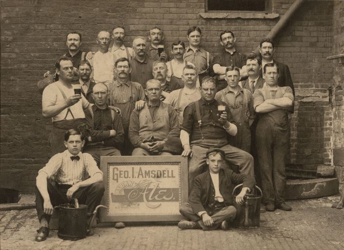 Group of men pose in front of a brick wall. Some are holding pints of beer. 